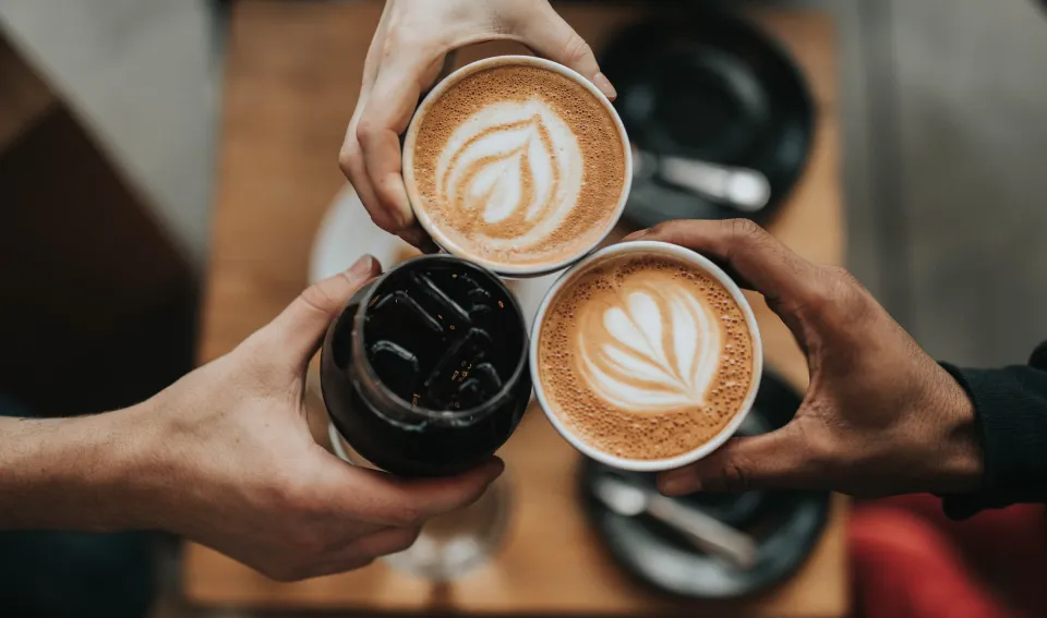 Three people raising latte-art coffees and an iced coffee over a café table